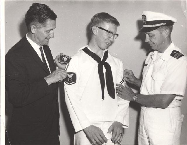 A man in a suit and a naval officer pin patches on a smiling sailor in uniform, marking a ceremony or achievement.