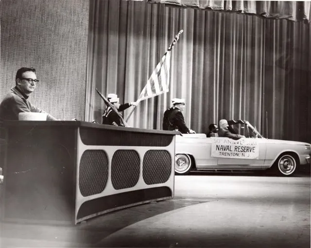 A man sits at a desk while a convertible with "Naval Reserve Trenton, N.J." banner passes by. Two sailors carry an American flag beside the car on a stage with a curtain backdrop.