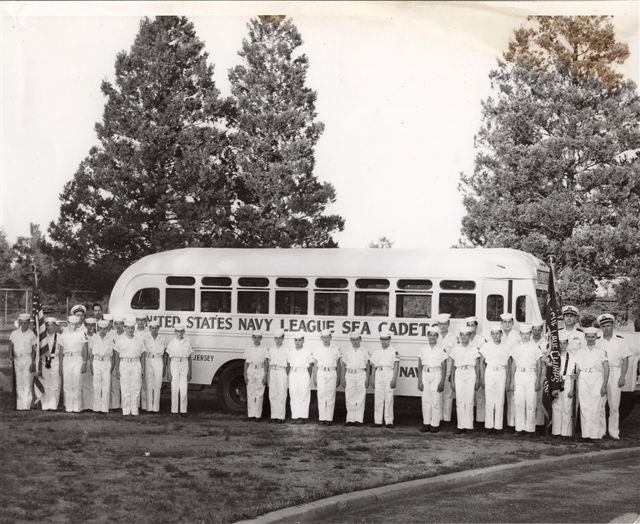 A group of cadets in uniform stands in front of a bus labeled "United States Navy League SFA Cadet." Trees and greenery are in the background.